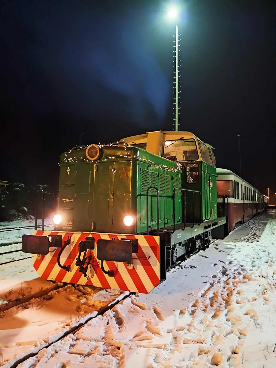 Osvětlená zelená lokomotiva 710.866 s historickým vlakem stojí v noci na zasněžené trati.